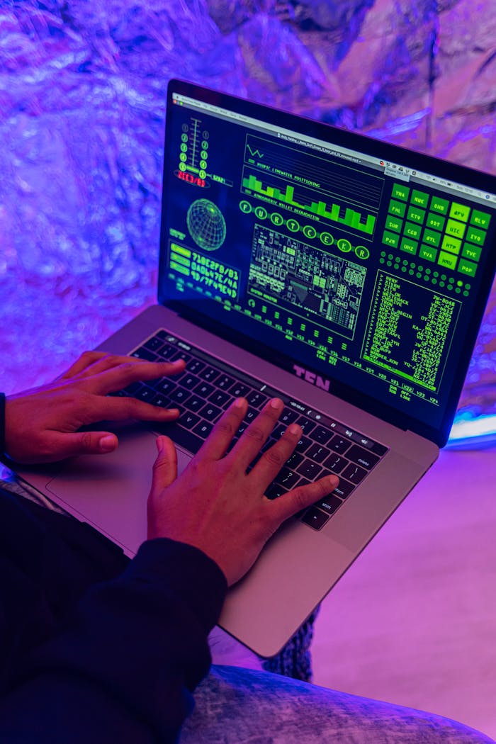 gallery-6 Close-up of hands typing on a laptop displaying green code, indoors, purple lighting.